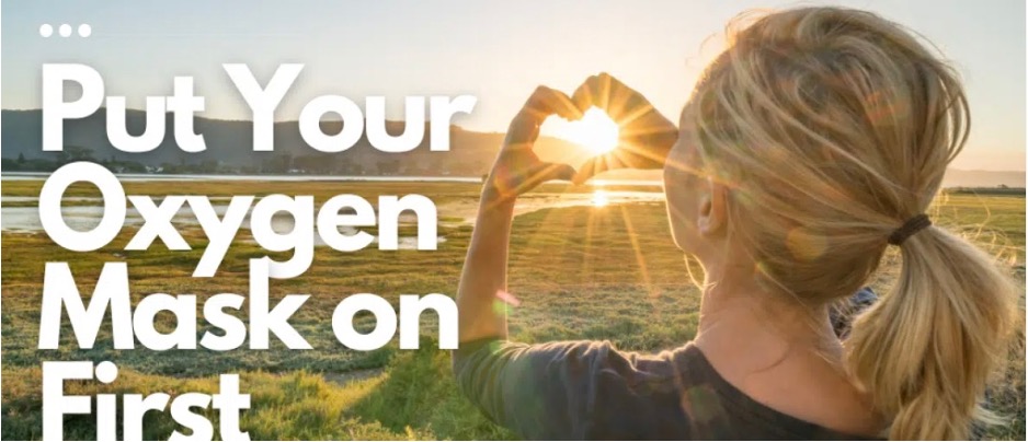 A woman standing outdoors in sunlight, making a heart shape with her hands toward the sun, with the text "Put Your Oxygen Mask on First" displayed prominently.